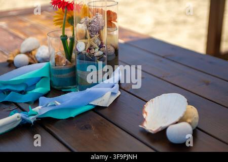 A coastal-themed table arrangement featuring tall glass cylinders filled with layers of sand, colorful pebbles, and seashells, accented by vibrant ger Stock Photo
