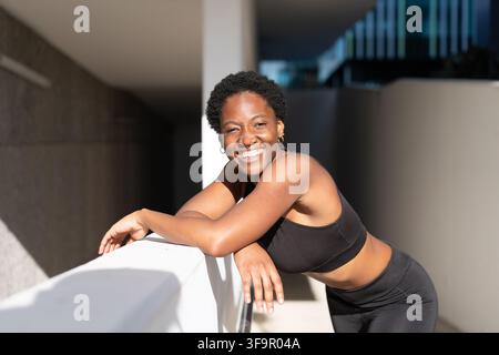 African American woman smiles while resting on a railing after a workout. Black woman looks happy and relaxed in modern urban surroundings. Stock Photo