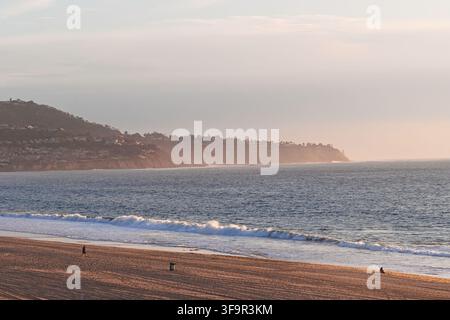 Beach scene at dusk with calm ocean waves. People relax on sand near water's edge. Sky has soft gray clouds above. Silhouettes show peaceful, contempl Stock Photo