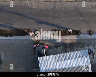 Aerial view of concrete road construction with workers using shovels to spread gravel along the roadside near a building in Kecamatan Manisrenggo, Jaw Stock Photo