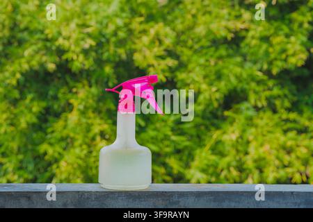 White spray bottle with a bright pink trigger nozzle sitting on a concrete ledge against a blurred green foliage background. Stock Photo