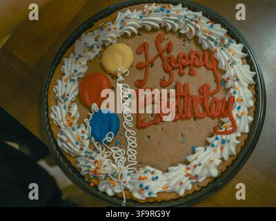 Top view of a giant frosted chocolate chip birthday cookie decorated with balloons and colorful sprinkles on a wooden table Stock Photo