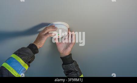 electrician in a safety uniform installing a white smoke alarm sensor on the ceiling. Concept of home fire safety, smoke maintenance, and security sys Stock Photo