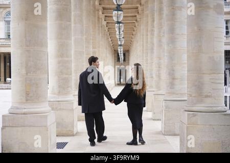 A stylish couple dressed in black walks hand in hand through the elegant colonnade of Palais-Royal in Paris, embracing romance and timeless architectu Stock Photo