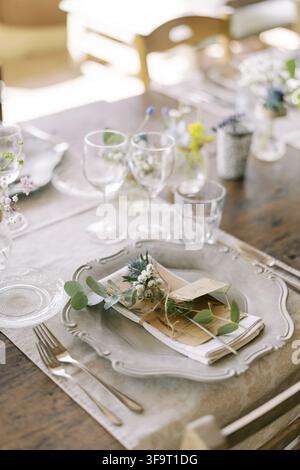 Close-up of a beautifully arranged wedding dinner table featuring a decorative plate, napkin with greenery, handwritten place card, wine glasses, and Stock Photo