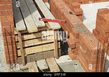 Workers are constructing a wall with clay bricks at a building site. A level tool is used to ensure proper alignment, while materials are organized ne Stock Photo