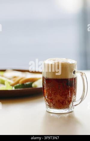 Beer cup standing near appetizers plate. Dark beer with thick high foam. Standing on smooth wooden surface of restaurant's or pub table. Looking mouth Stock Photo