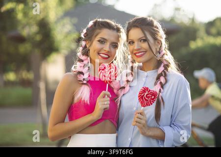 Pretty girlfriends with colorful braids holding candy heart on stick like heart and happy smiling at camera. Gorgeous and stylish women walking at str Stock Photo