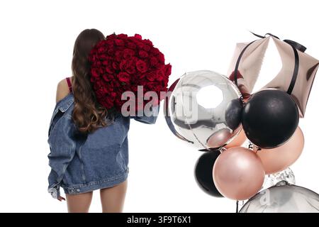 Woman's back in blue jeans jacket holding big bouquet of roses on her shoulder. Brunette girl with long hair getting presents on birthday. Stylish lad Stock Photo
