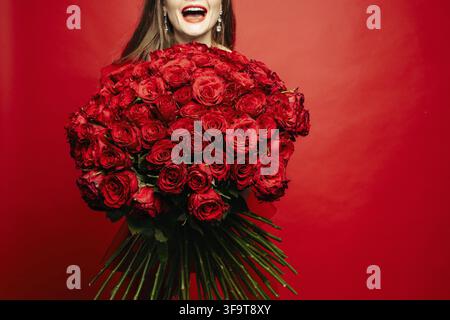 Selective focus of beautiful bouquet of red roses in hands of happy woman. Young lady laughing and keeping wonderful flowers on red isolated backgroun Stock Photo