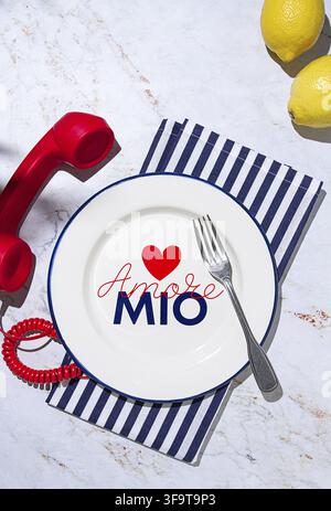 A round white plate sits on a striped napkin in a kitchen. It has the words Amore Mio written on it. A red phone and two lemons are placed nearby Stock Photo