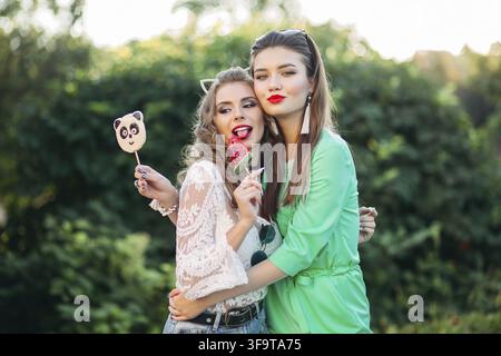 Two pretty and fashionable girlfriends holding candy on stick panda and water melon and happy smiling at camera. Gorgeous and stylish women walking at Stock Photo
