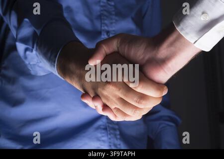 Low angle view of two businessmen hands shaking over dark gray background and light source at bottom side Stock Photo