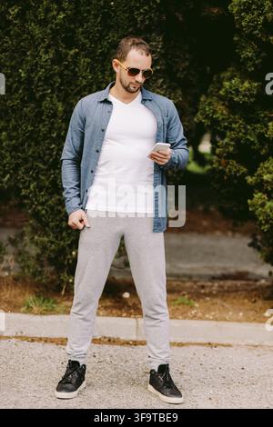 Man in white t-shirt holding phone mockup. Male wearing v collar white t shirt and using smartphone outdoors. Front view mockup Stock Photo