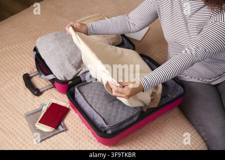 A women sits on the bed, folding a hoody while organizing a suitcase. Various clothes and travel accessories are neatly arranged inside the suitcase i Stock Photo