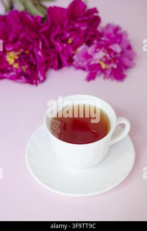 A cup of tea against the backdrop of a lush bouquet of burgundy peonies close-up on a pink background Stock Photo