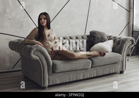 Full length of stunning brunette young model in beige laced romper and high heels posing on luxurious grey sofa with furry pillows Stock Photo