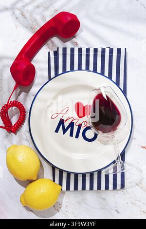 A round white plate sits on a striped napkin in a kitchen. It has the words Amore Mio written on it. A red phone and two lemons are placed nearby Stock Photo
