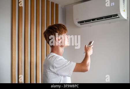 Young man adjusting air conditioner with remote control. Stock Photo