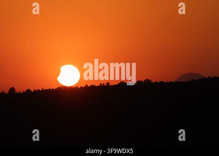 Beautiful landscape photo of a partial solar eclipse at sunset. The 'bitten' sun sets behind a dark silhouetted horizon of trees and mountains under a Stock Photo