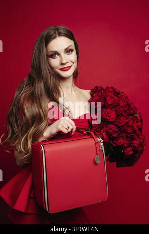 Fashionable studio portrait of stylish young woman with long brown hair in expensive red suit with leather red bag and bunch of red roses. She is smil Stock Photo