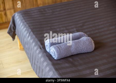 A gray bedspread with two rolled towels on neatly made bed, creating calm and organized bedroom setting with warm wood paneling and natural light Stock Photo