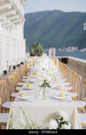 Long outdoor wedding dinner table with white flowers, lemons, and wooden chairs arranged on a terrace overlooking the sea and mountains at Heritage Gr Stock Photo