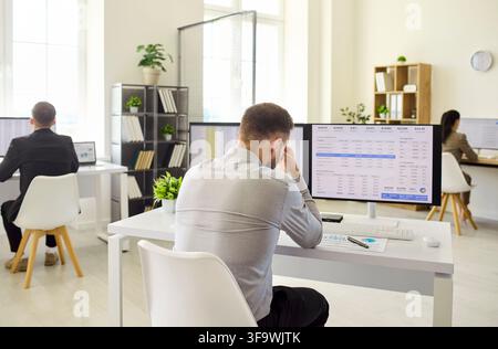 Stressed analyst man at office desk with finance data on monitors and spreadsheets Stock Photo