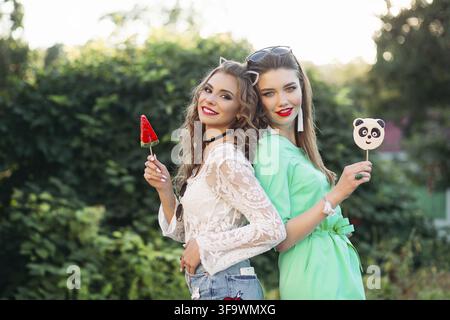 Two pretty and fashionable girlfriends holding candy on stick panda and water melon and happy smiling at camera. Gorgeous and stylish women walking at Stock Photo