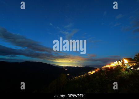 A mountain village at twilight with glowing hillside homes, dramatic clouds, dark valley, and distant city lights beneath deep blue sky Stock Photo