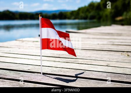 Austrian flag with red white red stripes stands on a wooden pier at a sunny lake. Gentle breeze moves the fabric and shadow. Clean alpine summer scene Stock Photo
