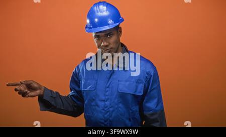 Man in blue hardhat and blue coveralls points finger to left, visible hand and finger, wearing safety helmet and work uniform in studio; focus guidanc Stock Photo