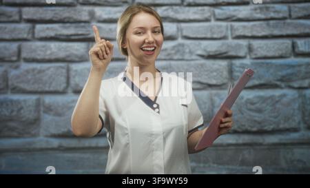 Woman nurse in white uniform pointing index finger and holding pink clipboard in studio; cheerful guidance. Stock Photo