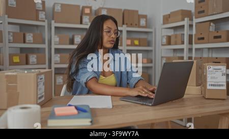 Woman typing on laptop and handling shipping papers at packing desk in warehouse storage room; entrepreneurial focus. Stock Photo
