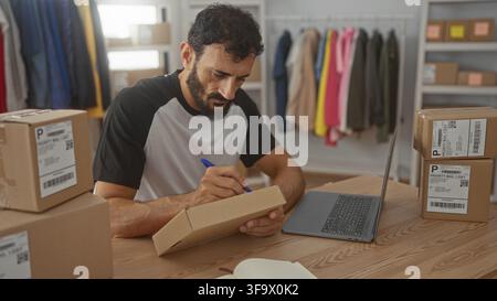 Man writing address on parcel with pen at packing table in studio surrounded by stacked shipping boxes, laptop and clothing rack; focused productivity Stock Photo