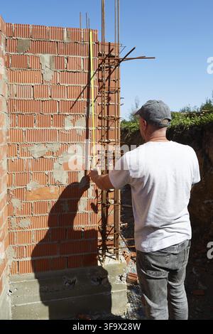A construction worker is measuring a brick wall at a building site, carefully checking the vertical alignment of bricks on a sunny day, ensuring quali Stock Photo