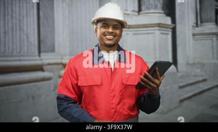 Hispanic man construction worker in hard hat points finger to tablet at building while checking plans in red jacket; confidence efficiency safety. Stock Photo