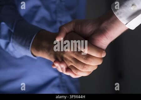Low angle view of two businessmen hands shaking over dark gray background and light source at bottom side Stock Photo