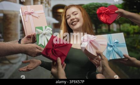 Redhead woman smiling and receiving wrapped gift boxes from multiple hands on a street, hands offering heart box and ribboned parcels; joy celebration Stock Photo