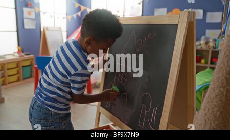 African american boy erases a chalkboard with hand while drawing a house on an easel in a daycare building filled with toys and bunting; playful focus Stock Photo