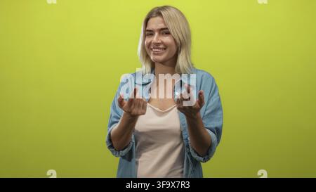 Woman beckons with fingers and open hands, smiling in a denim shirt in a green studio; friendly invitation joy. Stock Photo