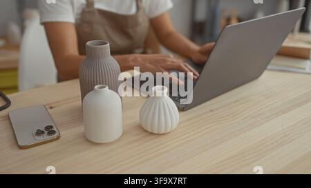 Woman typing on laptop showing hands beside three ceramic vases on wooden table in studio; focus craft calm. Stock Photo