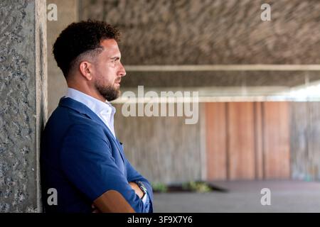 Professional businessman wearing a blue blazer standing with arms crossed and looking away while leaning against a concrete wall outside a modern offi Stock Photo