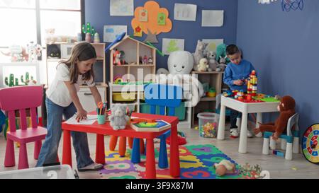 Girl leaning over a red table drawing with colored pencils while a boy stacks blocks at a small table in a colorful playroom; concentration. Stock Photo