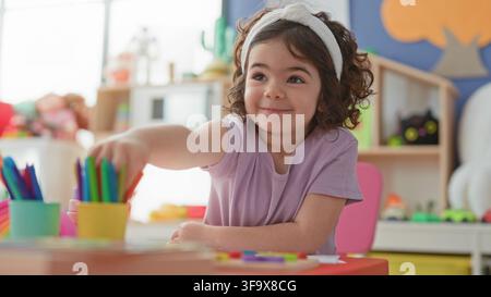 Girl with headband reaching for colored pencils on table among toys and shelves in bright preschool classroom; creative joy. Stock Photo