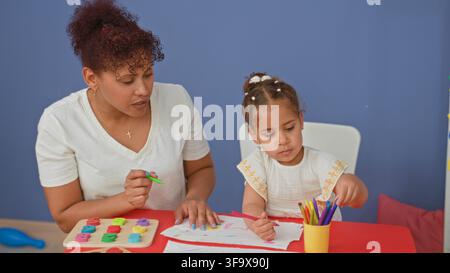 Woman guides girl's hand holding pink pencil as child draws on paper with colorful markers and wooden puzzle on red table in studio; nurturing learnin Stock Photo