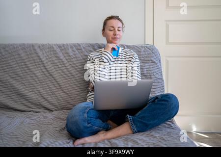 Thoughtful woman thinking on purchasing goods at online marketplace with laptop and credit card. Stock Photo