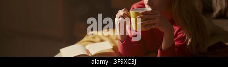 Caucasian young woman drinking coffee while lying on sofa with open notebook, relaxing at home. Image represents mental wellbeing and self care, suitable for lifestyle blog banner Stock Photo