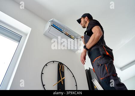 Air conditioner technician inspects an open wall mounted unit during maintenance at home. Male worker checks filters and internal cooling parts while Stock Photo