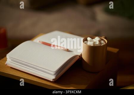 Open notebook with pen lying on pages next to mug of hot cocoa with marshmallows on wooden table. Image suitable for mental wellbeing, journaling, and cozy lifestyle marketing, close up Stock Photo